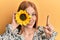 Young irish woman holding sunflower over eye smiling with an idea or question pointing finger with happy face, number one