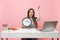 Young irritated woman spreading hands holding alarm clock sit, work at office with pc laptop isolated on pastel pink