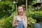 Young jocund woman talking by smartphone in garden near trees, wearing jeans sundress.