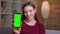 Young joyful brunette female shows upright green chroma screen of phone recommending the app at home.