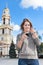 Young joyful woman a photograph with easily handled camera, looking at screen. Stands behind bell tower of church in Russia city