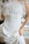 Young lady in luxurious white dress decorated with small, fluffy feathers stand indoor. Female lift fabric with hand.