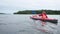 young lady rows bright orange sports kayak with little kid sailing along lake against forest and cloudy sky on nasty day