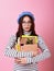 Young lady student in a stylish beret hat, with a cardboard box in her hands on a pink background