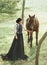 A young lady in a vintage dress strolls through the forest with her horse. The girl has a white blouse, a jabot, a tie