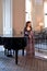 The young lady in a violet old-fashioned dress with a frill stands near the piano in the old interior of the mansion