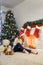 Young lady in white sweater sitting with cup near fireplace. New Year tree and home decoration. Candles and teddy bear, gifts are