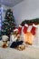 Young lady in white sweater sitting with cup near fireplace. new Year tree and home decoration. Candles and teddy bear, gifts are