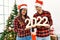 Young latin couple celebrating new year holding 2022 number standing by christmas tree at home