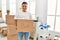 Young latin man smiling happy holding books cardboard box moving at new home