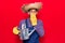 Young latin man wearing farmer hat and gloves holding watering can covering mouth with hand, shocked and afraid for mistake