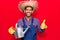 Young latin man wearing farmer hat and gloves holding watering can smiling happy and positive, thumb up doing excellent and