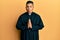 Young latin priest man praying with hands together making fish face with mouth and squinting eyes, crazy and comical