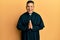 Young latin priest man praying with hands together smiling and laughing hard out loud because funny crazy joke