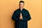 Young latin priest man praying with hands together winking looking at the camera with sexy expression, cheerful and happy face