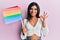 Young latin transsexual transgender woman holding rainbow lgbt flag doing ok sign with fingers, smiling friendly gesturing