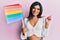 Young latin transsexual transgender woman holding rainbow lgbt flag smiling happy pointing with hand and finger to the side