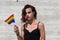 Young latin and transsexual woman on grey background holding the gay pride flag with her hand. Concept diversity, transgender, and