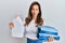Young latin woman holding jeans for laundry and detergent bottle making fish face with mouth and squinting eyes, crazy and comical