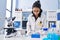 Young latin woman scientist writing on notebook holding pills at laboratory