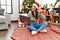 Young latin woman using laptop sitting by christmas tree waiving saying hello happy and smiling, friendly welcome gesture