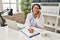 Young latin woman wearing doctor uniform talking on the telephone write on clipboard at clinic