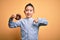 Young little boy kid eating unhealthy chocolate doughnut over isolated yellow background with surprise face pointing finger to