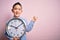 Young little boy kid holding big minute clock over isolated pink background very happy pointing with hand and finger to the side