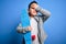 Young little boy kid skateboarder holding modern skateboard over blue isolated background stressed with hand on head, shocked with