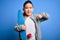 Young little boy kid skateboarder holding modern skateboard over blue isolated background with surprise face pointing finger to