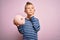 Young little caucasian kid banking money on piggy bank as wealth savings over pink background serious face thinking about