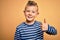Young little caucasian kid with blue eyes wearing nautical striped shirt over yellow background doing happy thumbs up gesture with