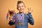 Young little caucasian kid wearing winner medals and victory award trophy over yellow background doing ok sign with fingers,