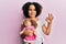 Young little girl with afro hair holding animal doll toy doing ok sign with fingers, smiling friendly gesturing excellent symbol