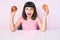 Young little girl with bang holding red apple and donut sitting on the table afraid and shocked with surprise and amazed