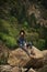 Young lonely tourist cutie girl sitting on stone on shore of river in mountains in wild terrain and satisfying thirst