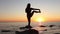 A young long-haired girl making yoga practice standing on a rock by the sea. Summer, sunset. Woman`s silhouette