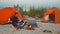 Young loving couple of tourists camping, sitting by the fire against orange tents on the beach