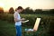 Young male artist, standing by easel, drawing portrait on canvas on green field in summer during sunset. Painting workshop in