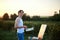 Young male artist, standing by easel, drawing portrait on canvas on green field in summer during sunset. Painting workshop in