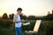 Young male artist, standing by easel, drawing portrait on canvas on green field in summer during sunset. Painting workshop in