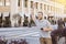 Young Male with a Big Smile Raises His Hand to a Rideshare Driver in the City - Warm Light in the Afternoon - Holding a Cell Phone