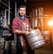 Young male brewer in leather apron holds barrel with craft beer at modern brewery factory