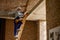 Young male builder in blue overalls and hard hat looking focused, climbing up the ladder while working at construction