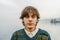 Young male with contemplative gaze, wearing sweater, against blurred lakeside backdrop