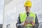 Young male engineer in reflector-vest and hardhat using digital tablet outside industry