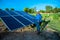 Young male engineer talking on phone with laptop in hand standing near solar panels, agriculture farm land with clear blue sky