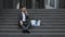 Young male jobless office worker in suit sitting on stairs outdoors. Fired man talking on phone with stuff in box