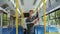 Young male passenger in medical face mask standing in aisle, using smartphone and holding handrail travels by trolleybus