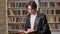 Young male student in black leather jacket standing in library and reading, book shelves background